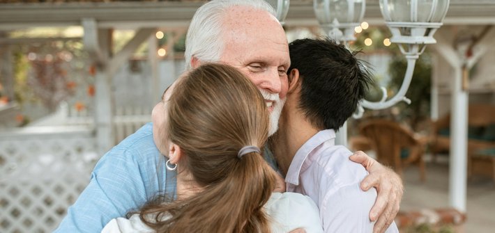 An elderly man hugs a younger man and woman