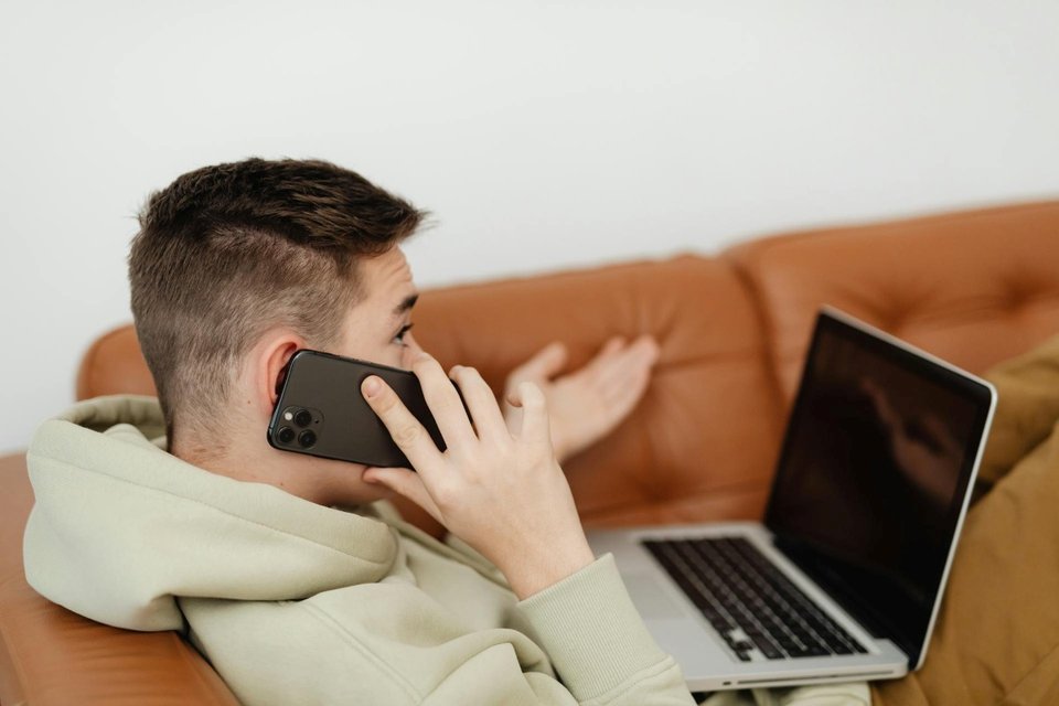 a teenage boy with a laptop on a sofa using his phone on a call