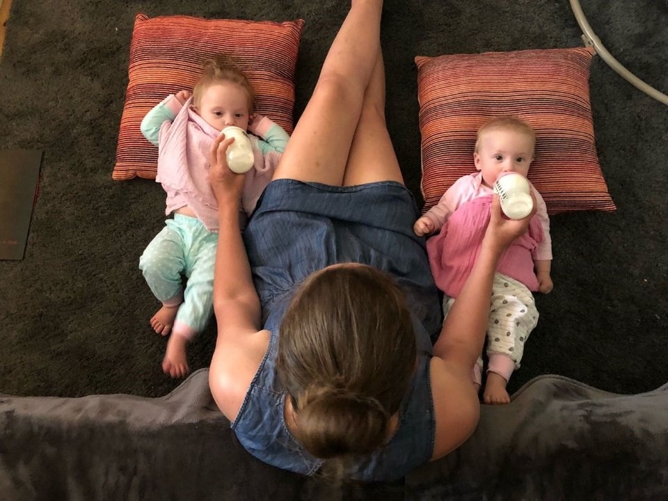 black woman tandem bottle feeding twin babies in bouncers