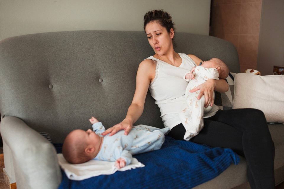 a woman holding a twin baby while comforting the other lying on the sofa