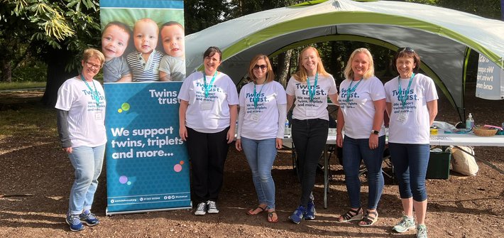 A group of people stand together in a park wearing Twins Trust t-shirts next to a Twins Trust banner