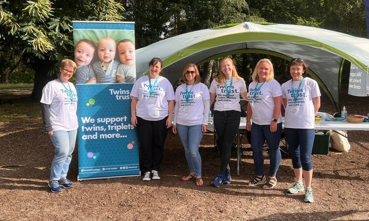 A group of people stand together in a park wearing Twins Trust t-shirts next to a Twins Trust banner
