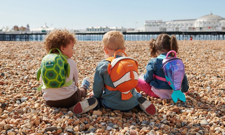 Three toddlers sit on a beach wearing different coloured backpacks