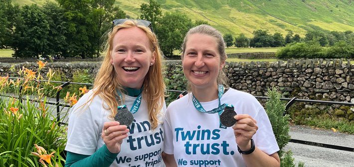Two women in Twins Trust t-shirts pose with medals in front of a background of green hills