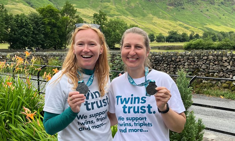Two women in Twins Trust t-shirts pose with medals in front of a background of green hills