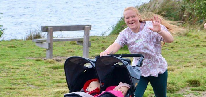 A woman waves at the camera as she runs pushing a double buggy