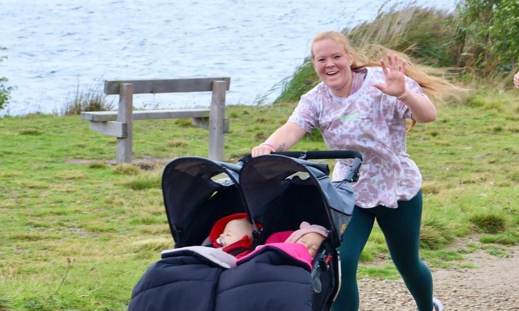 A woman waves at the camera as she runs pushing a double buggy