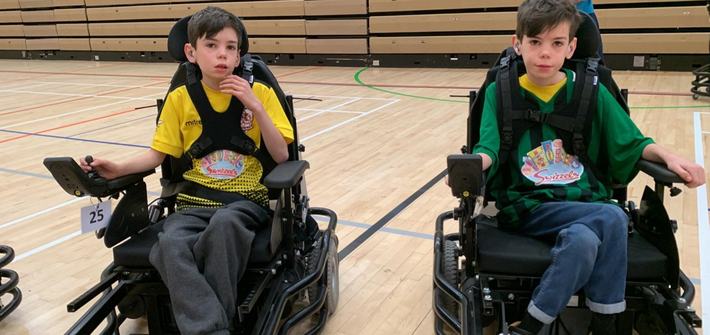 two boys sat in electric wheelchairs in a gymnasium