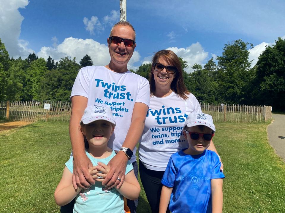 a father and mother wearing Twins Trust t-shirts and twin boy and girl both wearing Twins Trust caps in a park