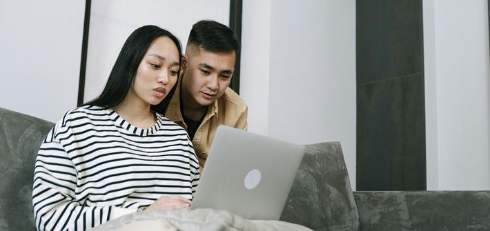 A woman sits on a sofa with a laptop on her lap as a man leans over her shoulder looking at the screen