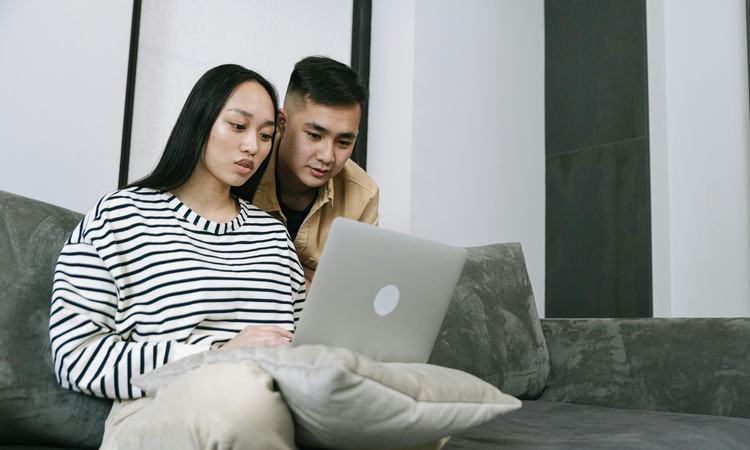 A woman sits on a sofa with a laptop on her lap as a man leans over her shoulder looking at the screen