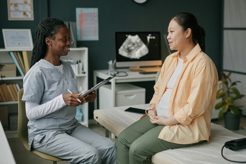 an obstetrician holding a clipboard speaking to a pregnant Asian woman