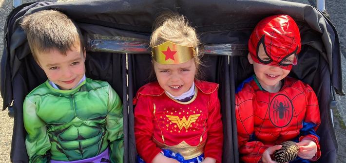 three children wearing superhero costumes sitting in a triplet pram