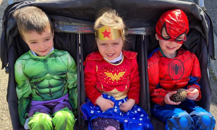 three children wearing superhero costumes sitting in a triplet pram