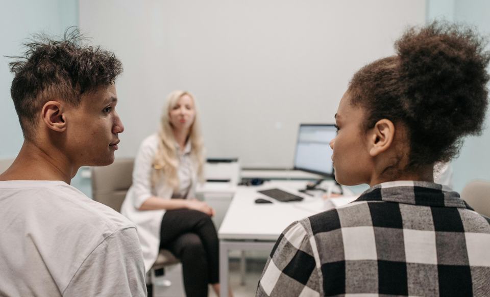 A young man and woman sit in the foreground with their backs to the camera looking at each other as a woman in a medical professional's jacket sits at a desk in the background