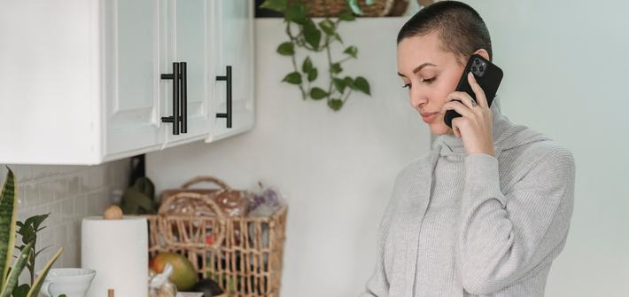 A woman stands in a kitchen with a phone to one ear making a cup of tea with the other hand