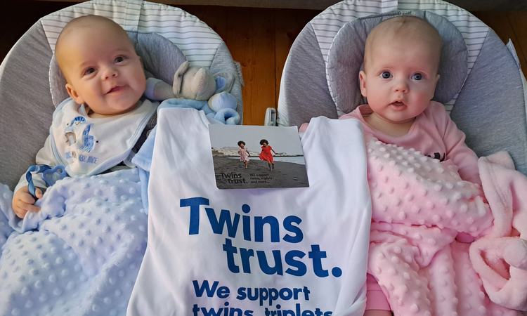 Twin babies lie in cribs with a Twins Trust bag between them