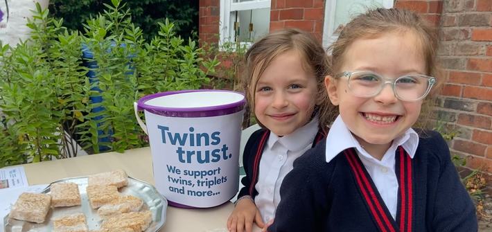 Two girls in school uniform stand next to a table with baked goods and a collection bucket on it