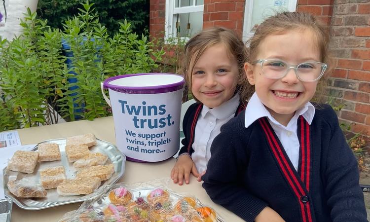 Two girls in school uniform stand next to a table with baked goods and a collection bucket on it