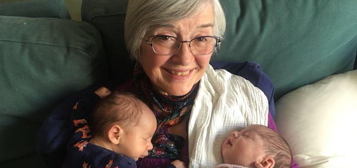 An older woman smiles at the camera as twin babies sleep on her chest