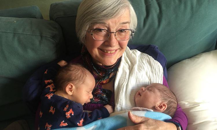 An older woman smiles at the camera as twin babies sleep on her chest