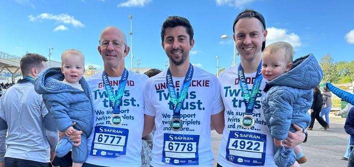 Three men stand in Twins Trust running shirts, two holding young twin boys