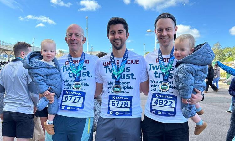 Three men stand in Twins Trust running shirts, two holding young twin boys