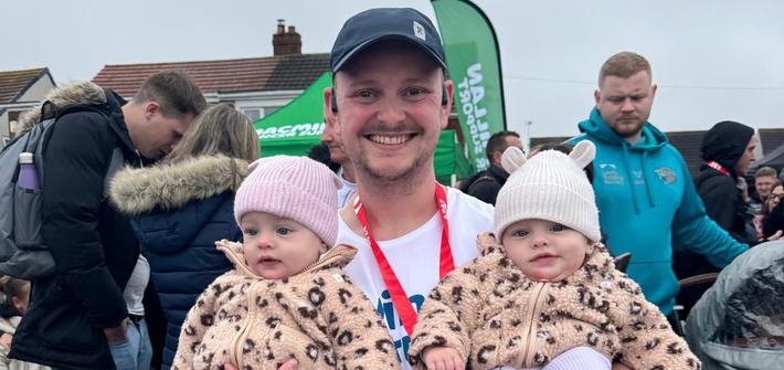 A man stands in a running shirt with a medal round his neck whilst holding twin babies