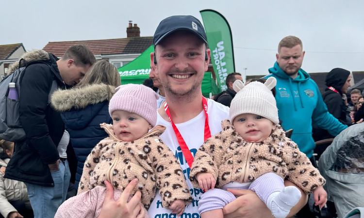 A man stands in a running shirt with a medal round his neck whilst holding twin babies
