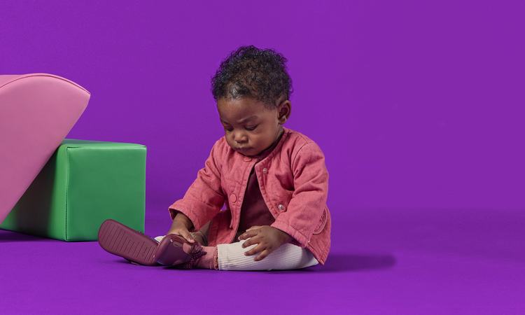 A toddler sits against a purple backdrop playing with their shoes