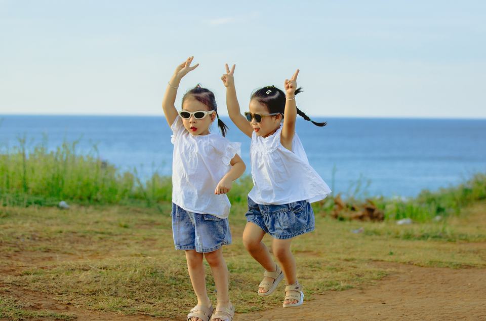 Young twin girls wearing sunglasses jump in the air with their hands up against a coastal background