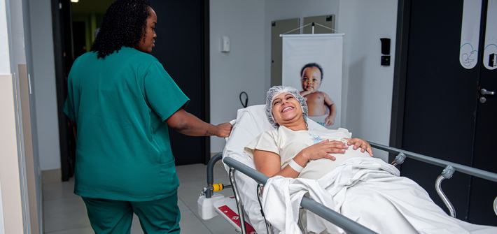 A pregnant woman is wheeled down a corridor on a hospital trolley with a nurse in scrubs walking beside her