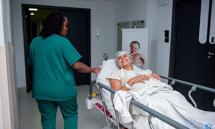 A pregnant woman is wheeled down a corridor on a hospital trolley with a nurse in scrubs walking beside her