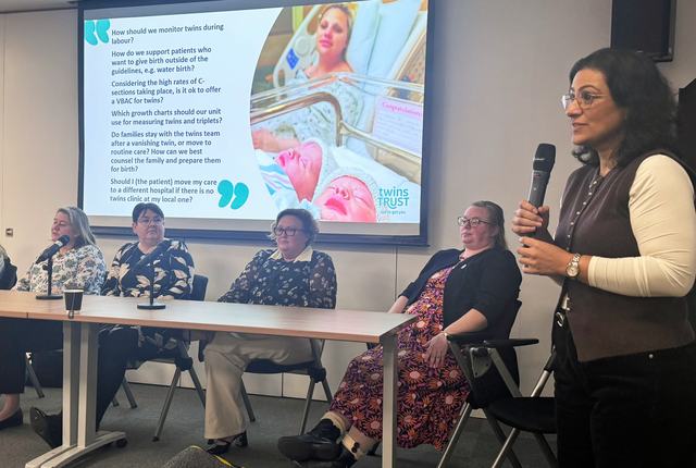 A woman stands holding a microphone to the right of a table with four women sat behind it