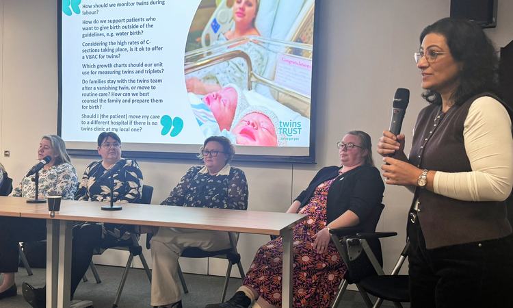 A woman stands holding a microphone to the right of a table with four women sat behind it