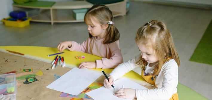 Two young girls sit at a table with pens laid out before them, drawing on sheets of paper