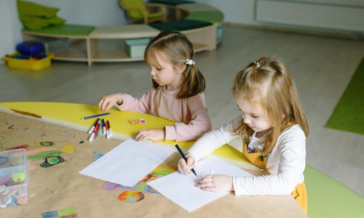 Two young girls sit at a table with pens laid out before them, drawing on sheets of paper