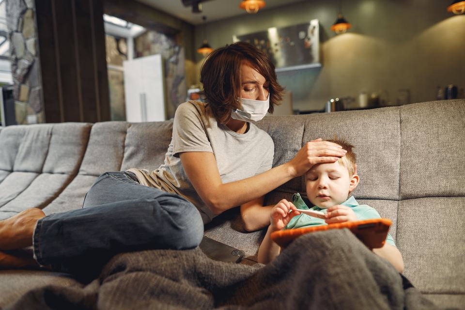 A concerned mother wearing a mask checks her child's temperature on the sofa