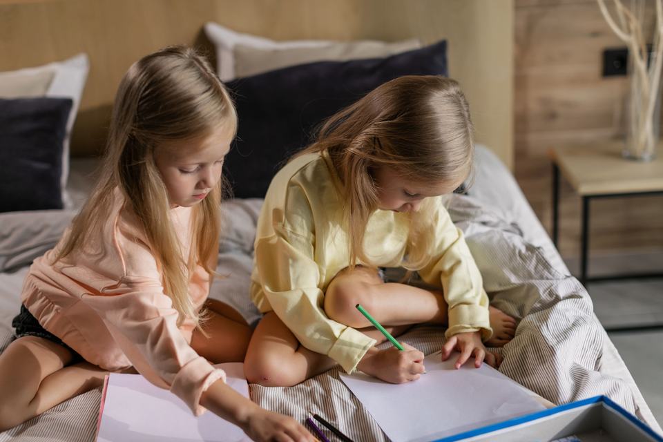 twin girls drawing with pencils while sitting on a bed