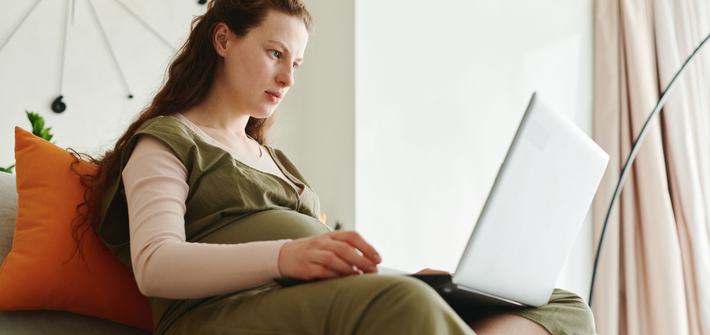 Pregnant woman working on a laptop in a cozy indoor setting, showcasing live online course