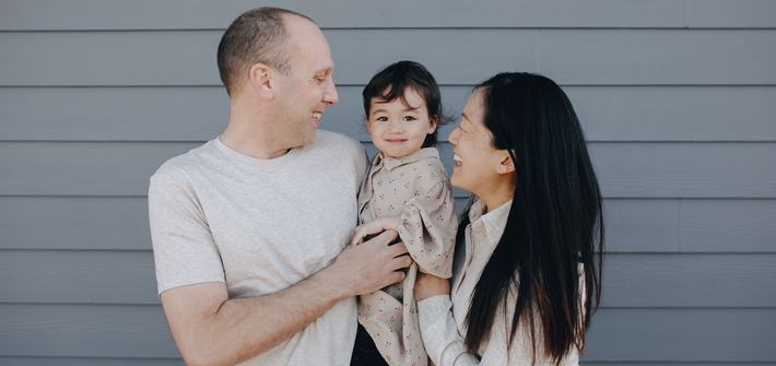 A joyful family portrait featuring a smiling child held by parents outdoors against a gray wall.