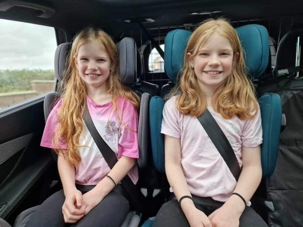 two pre-teen twin girls sat in high back car seats with seatbelts on in a family car