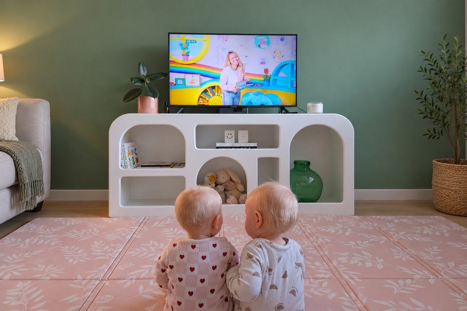two twin babies sat on a playmat watching their mother on TV