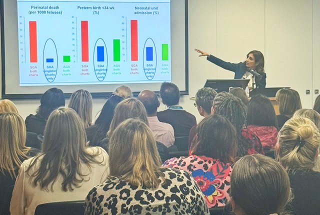 A group of people sit with their backs to the camera in the foreground, watching someone present in the distance