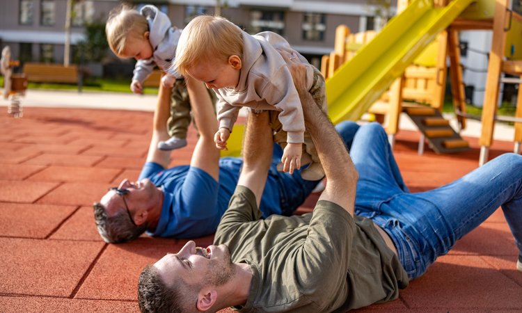 Two men lie on the ground with the playground in the background, each holding a baby in their arms above them