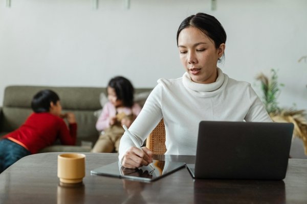 woman doing work on her tablet and laptop with two children in the background playing on the sofa